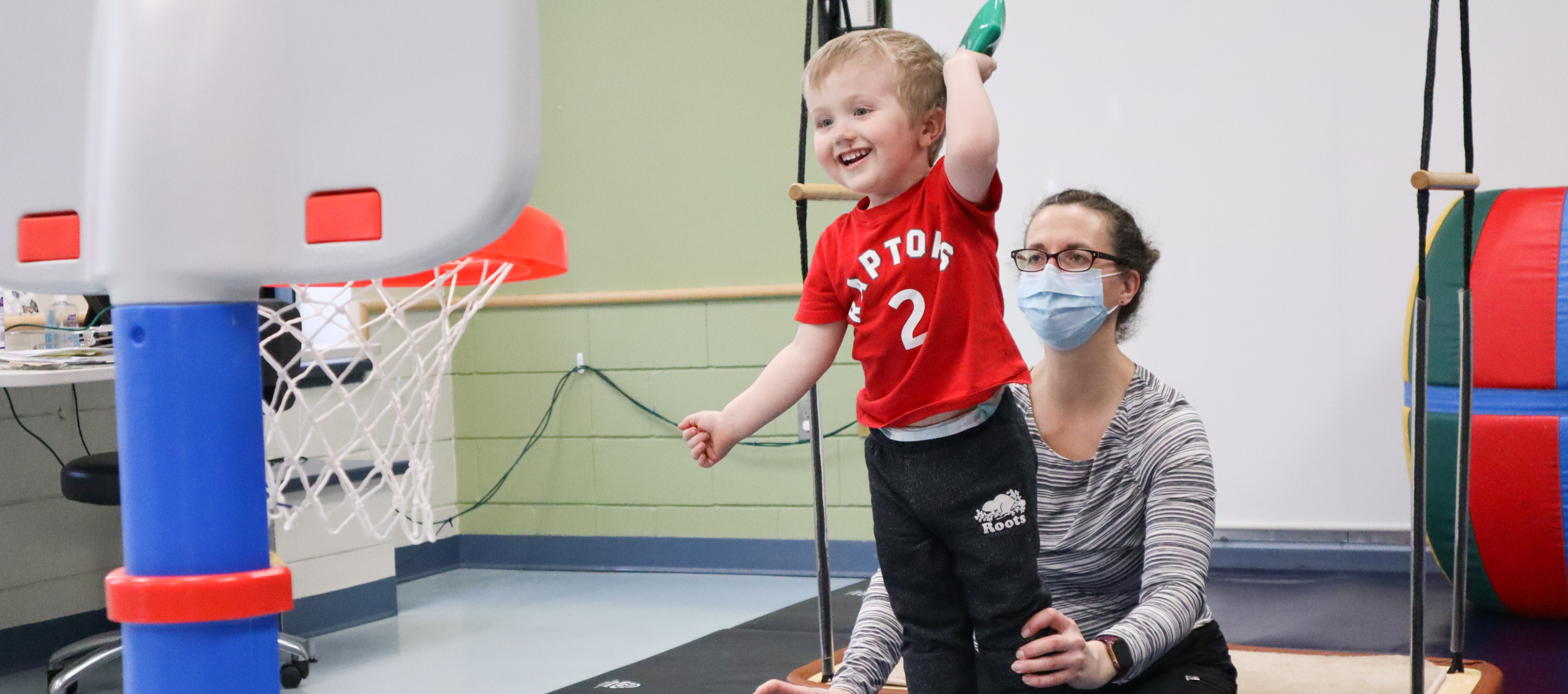Occupational therapist stabilizes male child as he throws object into children’s basketball net at N