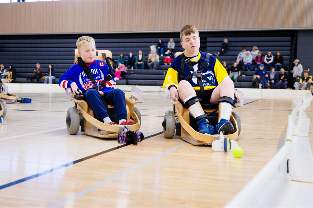 Two players in wooden chairs chase after a ball on a gym court